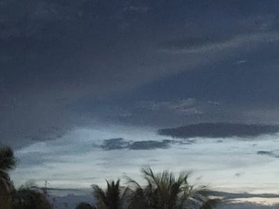 The image depicts a dark, cloudy sky with some lighter areas suggesting patches of light breaking through. In the foreground, the tops of palm trees are visible, indicating a tropical or subtropical environment. The overall impression is of an overcast or approaching storm. There are no discernible people, objects, or actions within the frame. The image is abstract and lacks specific details that would place it in Jelutung, Indonesia, or any other particular city.