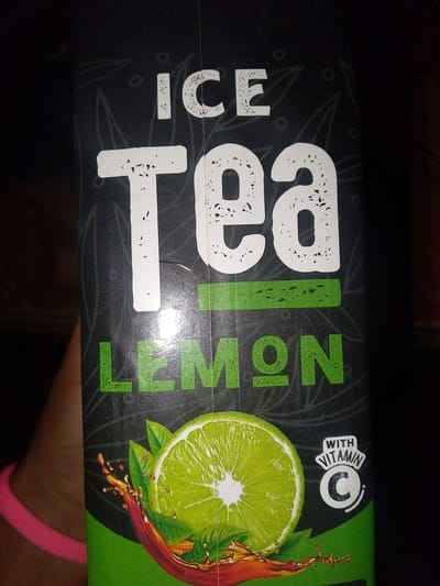 The image shows a close-up of a beverage carton for "ICE Tea LEMON". The carton has a dark background with stylized leaf designs. The words "ICE", "Tea", and "LEMON" are prominently displayed in large white and green letters. Below the text, there is a graphic of a sliced lime with liquid splashing around it. To the right of the lime, there is a white circular badge with the text "WITH VITAMIN C" and a large "C" inside. A person's arm and a pink bracelet are visible on the left side of the image. The lighting suggests the photo was taken indoors, possibly in a dimly lit environment. There is no information in the image that would allow for its location in El Maco, Venezuela.