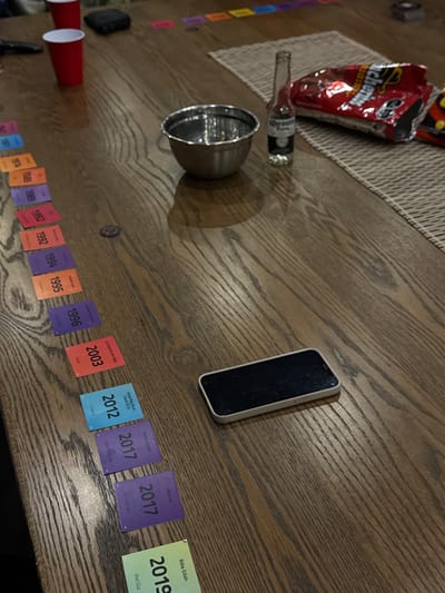 A top-down view reveals a cluttered wooden table. A bag of Takis, a Corona bottle, and a metal bowl sit near the left edge. To the right, a smartphone rests close to a timeline of cards with years and names like "Maluma" and "Billie Eilish."  The timeline spans from the 1970s to 2019. Red cups are visible in the lower-left corner, with miscellaneous objects scattered around the scene.