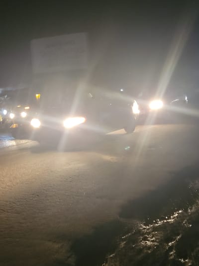 The image depicts a nighttime street scene in Kwamba, Nigeria, characterized by heavy fog or mist, which significantly obscures visibility and creates a diffused lighting effect. The foreground shows a wet, dark asphalt road, reflecting the bright headlights of vehicles. Several cars are visible, their headlights on and casting beams through the fog. One vehicle in the center has its headlights prominently illuminated. In the upper left quadrant, a partially obscured sign is visible, bearing text that appears to be an advertisement or business name, with lettering like "BOARD" and "GRAPHICS" discernible. The overall atmosphere is atmospheric and hazy, typical of foggy conditions at night in a city environment.