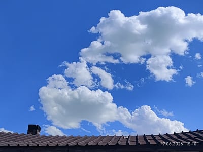 The image depicts a rooftop with a brown, corrugated tile surface under a bright blue sky filled with puffy white cumulus clouds. A dark, rectangular chimney or vent structure stands on the left side of the rooftop. The timestamp "17.06.2026 15:10" is visible in the lower right corner of the image. The scene is devoid of people and any discernible activity. The environment appears to be outdoors, possibly in a residential area of Üştöbe, Kazakhstan, given the context.