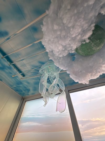 The image shows an indoor scene with a whimsical decoration hanging near a window. The decoration consists of white, fluffy material resembling clouds, a green, rounded object possibly representing a planet or landmass, and a translucent, iridescent ribbon sculpture hanging below the "clouds". The decoration is suspended by multiple white rods. The wall behind the decoration is tiled in a blue and white pattern. The window is partially visible, with a view of the sky at sunset or sunrise. The exact location in Novo-Nikol'skoe, Russia cannot be determined from the image. There are no people visible in the image.