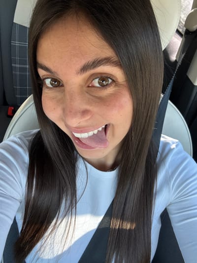 A female individual is depicted in a close-up, occupying the front passenger seat of a vehicle in Antibes, France. She has long, straight dark brown hair, dark brown eyes with visible reflections, prominent dark eyebrows, and fair skin with freckles across her nose and cheeks. She is wearing a white long-sleeved top. A black three-point seatbelt is fastened across her chest, extending from her left shoulder. The subject is looking directly at the camera with a wide smile, her mouth open and tongue fully extended. The vehicle's interior features a white headrest behind her and a dark grey checkered or plaid fabric on what appears to be a rear seat or door panel in the upper left background. Direct sunlight illuminates the scene, creating noticeable shadows.