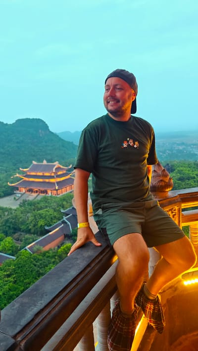 A man is perched on a railing overlooking a valley with a Buddhist temple. The man is wearing a dark green t-shirt with a cartoon graphic, green shorts, a black baseball cap, and plaid shoe covers. He is smiling and looking to his left. The temple, illuminated with warm lights, has a multi-tiered roof and is surrounded by lush green trees. In the background, mountains are visible under a clear, pale blue sky. The railing is made of dark wood with ornate carvings, and a bright yellow wristband is visible on the man's left wrist. The scene is likely in the vicinity of Tây Hoa Lư Ward, Vietnam, given the architectural style of the temple and the surrounding landscape.