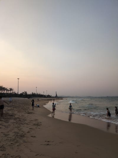 The image depicts a beach scene at sunset in Tuy Hòa, Vietnam. The sky is a gradient from light grey to orange near the horizon. The beach is sandy and textured with numerous footprints. Several people are present, some wading in the water at the edge of the waves, others standing or walking along the shore. Streetlights are visible along the beach. The water is slightly choppy with small waves.