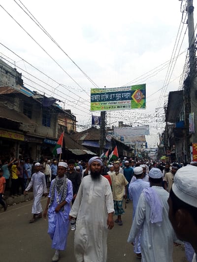 The image depicts a crowd of people walking down a street in the city of Bhola, Bangladesh, specifically at the coordinates 22.686 latitude and 90.644 longitude. The majority of the people are dressed in traditional white clothing, many with beards and some wearing white skull caps. They are likely participating in a religious procession, as evidenced by the numerous banners and flags, one of which features the inscription "মাঝারপাড়া মসজিদ ও মাদ্রাসা" ("Madrassa and Mosque of Madhyapara"). The street is lined with buildings, some with shops and signs, and power lines are strung overhead. There are many people on the street, some walking in groups and some individually. Several people are holding flags and banners.