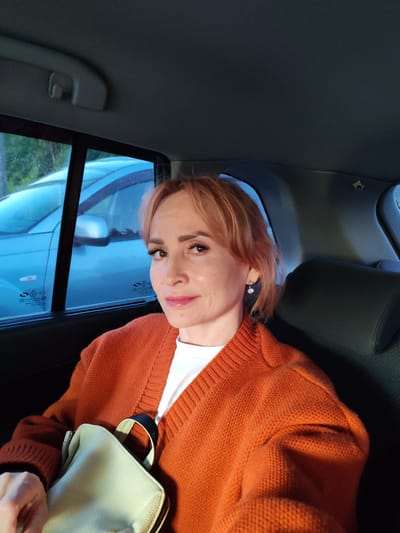 A woman with blonde hair, wearing an orange cardigan and a white t-shirt, is sitting in the back seat of a car. She is holding a light-colored purse on her lap. The window behind her shows a blue car and trees outside. The scene appears to be outdoors, possibly in the city of Bol'shie Mkhi, Russia, given the context. The woman is looking towards the camera and smiling slightly. The lighting suggests it is either daytime or dusk.