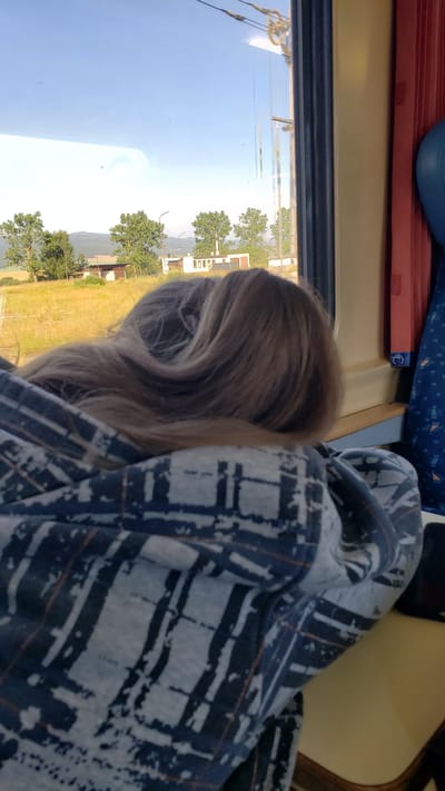 A person with long, blonde hair is resting their head on their arm and shoulder, wrapped in a blue and white patterned blanket. They are looking out of a train window at a rural landscape under a clear blue sky. The landscape features dry, yellowish grass, several green trees, and a few small, light-colored buildings, likely in Vydrník, Slovakia. The train car's interior is visible, with a yellow wall and a glimpse of a blue patterned seat or fabric. The lighting suggests it is daytime, possibly late afternoon.