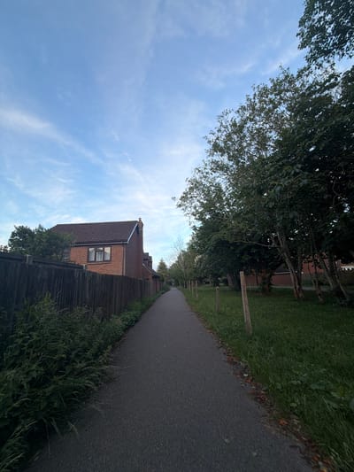 The image displays a straight, asphalt footpath or cycle path extending into the distance under a light blue sky dotted with scattered thin white clouds. This scene is located in a suburban residential area of Maidstone, United Kingdom.
On the left side of the path, a dark, weathered wooden fence runs parallel to the path. Behind this fence, a red-brick house with a dark tiled roof is partially visible, featuring a prominent brick chimney on its right side and a window with leaded glass and internal curtains. Dense green foliage, including overgrown weeds and shrubs, lines the base of the fence and slightly encroaches onto the path.
On the right side of the path, a strip of well-maintained green grass is separated from the path by a series of evenly spaced, short wooden posts. Beyond the grass, a line of mature deciduous trees with full green leaves stands, partially obscuring further brick houses in the distance. The lighting suggests either early morning or late afternoon, with soft shadows and illumination. No people or animals are visible, indicating a quiet environment.
