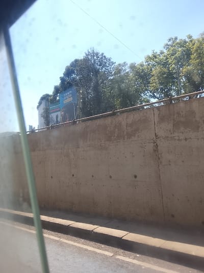 The image, captured from a moving vehicle, presents a roadside scene under a clear blue sky. In the immediate foreground on the left, a blurred, vertically oriented green or gray element partially obscures the view, suggesting a car window frame or pillar. To its right, a section of asphalt road is visible, marked by faint white lines. Adjacent to the road is a concrete curb, leading to a textured, light brown concrete retaining wall that dominates the midground. The wall appears aged and has visible seams and minor irregularities. A simple metal railing runs along the top of this wall.
Above and behind the wall, a dense canopy of green trees stretches across the upper part of the image, with some lighter-colored foliage towards the right, indicating bright sunlight. Nestled among the trees and partially visible above the wall on the left side of the background is a large billboard. The billboard features a blue section at the top with white text that reads "IT'S UP TO YOU" and a number "9772" below it. Further down on the billboard, on a darker background, more text is partially legible, appearing to say "ONE COVID, ALL V..." or "STOP COVID, ALL V...". A silhouette of a person is also visible on this lower section of the billboard, suggesting a public awareness campaign.
The bright, unfiltered sunlight suggests it is daytime, likely mid-morning or early afternoon, with no clouds in the sky. No people are explicitly visible in the scene, only the stylized figure on the billboard. The overall impression is an urban or suburban setting, possibly in Kiambu Township ward, Kenya.