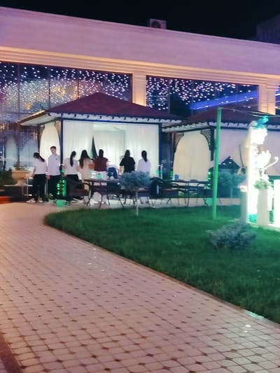 The image captures an outdoor seating area of a restaurant or cafe in Aksu, Kazakhstan, at night. A paved walkway with a diamond pattern leads the eye towards a covered seating area with white drapes. Several individuals, appearing to be staff and patrons, are visible within and around the seating area. Some are standing, while others are seated at tables. The building behind the seating area is illuminated with numerous small white lights against a dark background, suggesting a festive or decorative lighting scheme. A well-maintained green lawn is situated between the walkway and the seating area. The overall scene depicts a nighttime social gathering at an establishment.