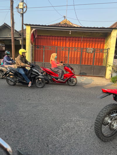 In Sugih Waras, Indonesia, on a paved road, three people are riding motorcycles. A man wearing a yellow helmet and a brown jacket is riding a black motorcycle in the foreground, with a black backpack attached to the back of the motorcycle. To his left, another person wearing a blue jacket and a black helmet rides a dark motorcycle. To the man's right, a woman in a pink shirt, orange headscarf, and patterned pants rides a red motorcycle, wearing a light blue mask. Behind them, a yellow building with an orange metal gate is visible, and power lines run overhead. The sun casts shadows on the road, suggesting it is late afternoon. A portion of another red motorcycle's wheel is visible in the lower right corner of the frame.