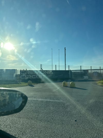 The image depicts the entrance to the Cremona Circuit in San Martino del Lago, Italy. The sun is shining brightly, casting lens flares across the scene. In the foreground, a portion of a car's side mirror is visible on the left. The road leads towards a large, dark structure with "CREMONA CIRCUIT" emblazoned on it in large, silver letters. Below this, smaller, cursive text reads "Angelo Bergamonte". Two yellow bollards are positioned in the middle of the road. Behind the structure, a fence and what appear to be flagpoles are visible against a clear blue sky. The overall environment suggests an outdoor, possibly racing-related facility.