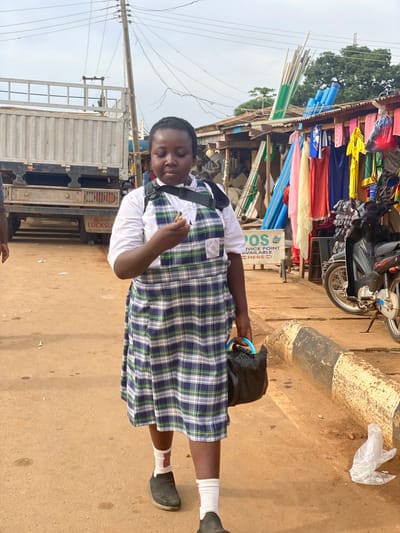 A girl wearing a school uniform is standing on a dirt road in Kafanchan B, Nigeria. The girl is wearing a plaid blue and green dress with a white shirt underneath, white socks, and black shoes. She is also wearing a black backpack and carrying a black plastic bag with a blue handle. She appears to be eating. Behind her is a truck and a shop selling building materials such as pipes and fabrics, and a motorcycle parked nearby. A sign says "POS SERVICE POINT AVAILABLE HERE". There is trash on the ground.