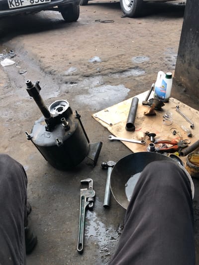 A mechanic's workspace shows scattered tools and car parts on a concrete floor. A large, black, cylindrical car part with a protruding pipe dominates the scene. Beside it, a large wrench and other metal components are visible. A white container sits atop a piece of cloth, surrounded by smaller tools like wrenches and pliers. The bottom left corner features a car's license plate. A person wearing gray pants and black shoes stands nearby.