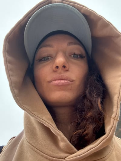 A medium-angle shot captures a woman's face framed by a light brown hooded sweatshirt and a light gray baseball cap. The woman has fair skin, brown hair, and green eyes. Her gaze is directed towards the viewer. The image is shot from a low perspective, looking up at the woman. It is difficult to determine the location without more information, and Binh Lu, Vietnam is not apparent in the image.