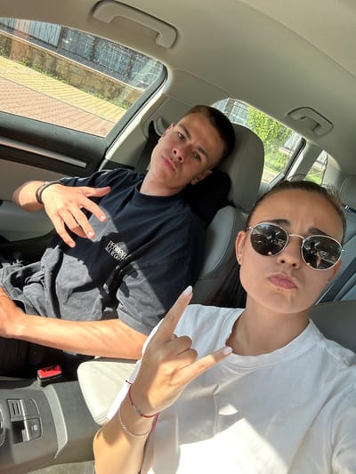 Two young people are seated inside a car, a selfie is taken. The man in the driver's seat, is wearing a black t-shirt, and has his left hand with a bracelet around his wrist, resting on his lap. The woman in the passenger seat, is wearing a white t-shirt and sunglasses. She is making a hand gesture with her right hand, and looking towards the camera. The background shows a residential area with buildings and vegetation. The location is identified as Vrachesh, Bulgaria.
