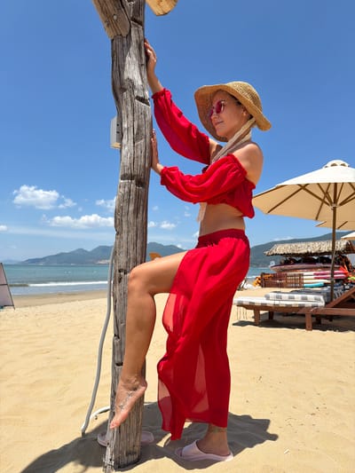 A woman in a red off-the-shoulder top and a flowing red skirt stands on a sandy beach, leaning against a weathered wooden post. She is wearing a straw hat, sunglasses, and light-colored slides. Her left leg is bent, with her foot resting on the post. She has a tattoo on her left ankle and a small navel piercing. In the background, the ocean meets the shore, with distant mountains under a clear blue sky with scattered clouds. Beach umbrellas and lounge chairs are visible further down the beach. This scene is likely located in Quy Nhon Ward, Vietnam, a coastal city known for its beaches and scenic views.
