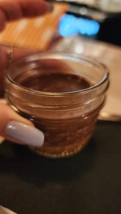 A human hand holds a small, clear glass jar containing a dark brown, opaque substance that fills approximately two-thirds of the container. The visible fingernails on the hand are coated with light lavender nail polish. The jar rests on a dark, flat surface. The background is significantly out of focus, displaying a prominent, blurred bright blue light source in the upper-right quadrant, adjacent to a less defined orange-tan rectangular object. The environment appears to be an indoor setting with dim, ambient lighting. The precise location within the city of Jackson, United States of America, cannot be determined from the visual information provided.