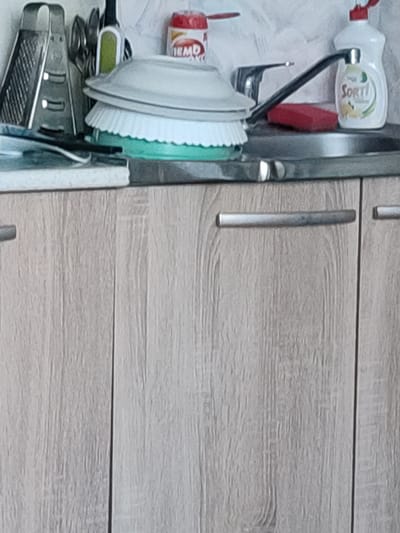 The image depicts a kitchen sink area with wooden cabinetry. Visible objects include a grater, various utensils, a red cleaning sponge, a bottle of "Sorti" dish soap, and a red can of "EMO" product. A stack of plates is also present, along with a kitchen faucet and sink basin. The scene appears to be indoors, with the lighting suggesting it is daytime. The cabinetry has a light wood grain pattern. The overall setting is a domestic kitchen. No people are visible in the image.