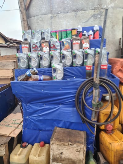 The image depicts an outdoor storage area, likely in Nyanya, Nigeria, characterized by a rough concrete floor and overhead structures that suggest a covered but open-air space. The primary subjects are a collection of various containers, predominantly plastic bottles filled with liquids, stacked on top of each other. These include numerous gray bottles of what appears to be engine oil, some with red and white labels, and several brown and green bottles. Interspersed among these are a few larger, yellow plastic jerrycans with red caps, also stacked.
A prominent object is a black coiled hose, seemingly industrial, draped over a metal rod that runs horizontally across the frame. This rod, bound with tape and some green material, supports the hose and also passes over some of the stacked bottles. The bottles and jerrycans are arranged on various surfaces, including wooden planks and a large, vibrant blue tarp that covers a significant portion of the stacked items.
In the background, other industrial elements are visible, such as orange barriers and more yellow jerrycans. A portion of a cardboard box with visible text is also present in the lower right. The lighting suggests it is daytime, with natural light illuminating the scene. The overall impression is one of a disorganized but functional storage area for liquids, possibly for commercial or industrial use. No people are visible in the image. The visible text on the cardboard box appears to read "AMP AMMO".