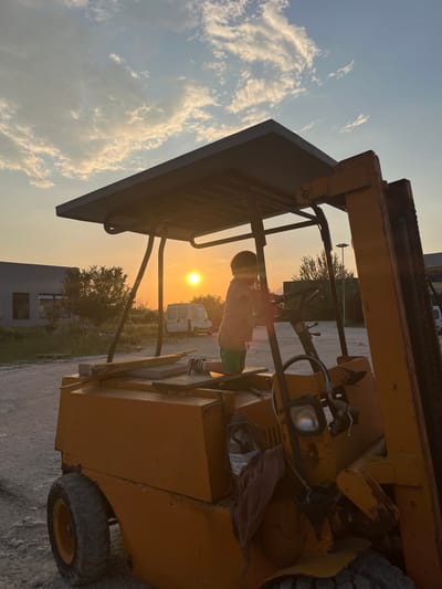 A child is standing on the seat of a yellow forklift, silhouetted against the setting sun. The sky is pale blue with white clouds on the left side of the frame. The forklift is parked on a gravel surface in Pobrdje, Montenegro. In the background, there are trees, a white van, and a building with a dark gray facade.