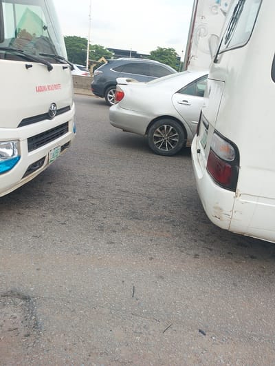 The image depicts a traffic scene on a road in Dutsen Alhaji, Nigeria. The foreground is dominated by the front and side of a white bus, with the text "KADUNA ROAD ROUTE" prominently displayed in red. The bus has a Toyota logo visible on its grille and a license plate that reads "224 JGS 6." The front bumper has blue accents.
To the right of the white bus, another white vehicle, likely a minibus or a van, is partially visible, showing its rear section with taillights and some scuff marks on the bumper.
In the background, a silver sedan is parked behind the white bus. Further back, a dark-colored SUV is also visible, along with lush green trees lining the roadside. Overhead, a utility pole with wires is present. The sky appears overcast, suggesting it might be a cloudy day. The road surface is asphalt, showing signs of wear and tear with small pebbles and cracks. The overall impression is that of a typical traffic situation in a Nigerian urban or suburban area. There are no people visible in the frame.