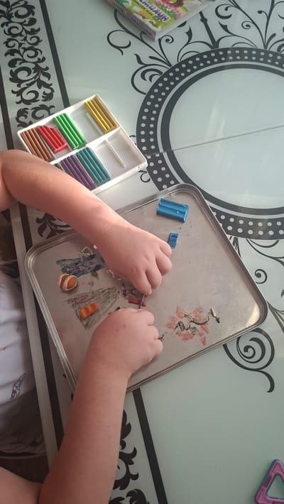 A high-angle shot shows a child's hands working with modeling clay on a metal tray. The child's arms and hands are the main subjects in the foreground. The child is manipulating small pieces of clay of various colors, including orange, blue, red, and purple. In the background, a white tray contains rows of unworked modeling clay sticks in colors such as brown, red, green, yellow, purple, and teal. A book or box with colorful graphics is partially visible in the upper right corner. The scene takes place on a table with a patterned surface, featuring decorative black swirls and circles. A portion of the table also has a circular design with polka dots. The overall environment appears to be indoors, possibly in a home setting. The location is stated to be in Voinova, Moldova.