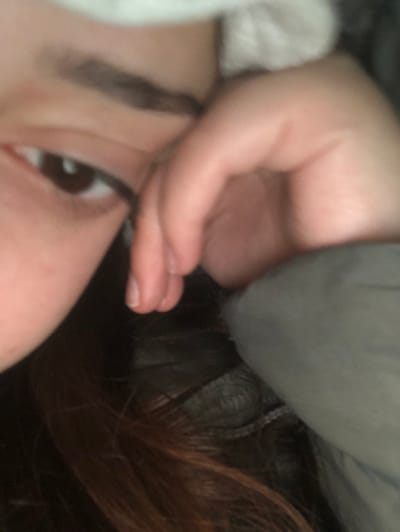 The image displays a close-up, rotated view of a person lying down, likely on their side. The perspective is rotated approximately 90 degrees counter-clockwise.
In the lower half of the frame (if viewed upright), the left side of a person's face is visible, including a prominent brown eye looking upwards, with distinct eyelashes and a well-defined eyebrow. The skin appears fair.
A hand, presumably belonging to the same person, is positioned in the upper center of the frame (if viewed upright), with its fingers gently curled and resting near the face, partially obscuring the cheek and temple area. The fingernails are visible. The hand's position suggests a relaxed posture, perhaps resting the head or shielding the eye.
To the right side of the frame (if viewed upright), a mass of dark brown or reddish-brown hair (possibly auburn) is visible, flowing downwards. Portions of the hair are covered or intermingled with a grey fabric, which appears to be a blanket, pillow, or an item of clothing like a hooded garment, occupying the upper right quadrant. Another lighter-colored, possibly white, soft fabric is vaguely visible on the far left edge of the image, likely part of clothing or bedding.
The overall scene suggests an indoor setting, possibly a bedroom or living area, with the person in a relaxed or resting pose. The lighting is soft and diffused, with no harsh shadows, implying a natural or indirect light source. There is no visible text in the image.