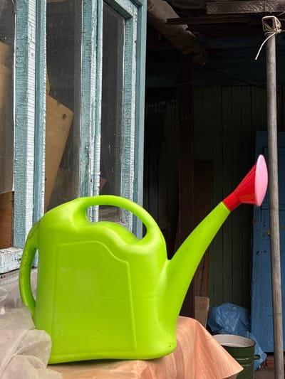 A neon green plastic watering can with a red and pink nozzle is positioned prominently in the foreground, resting on a light brown, textured surface that appears to be a draped cloth. The watering can occupies the majority of the frame. Behind it, a rustic, weathered wooden structure is visible. This structure features a window with peeling light blue paint and visible wooden planks. Beyond the window, the interior of the structure is dark and appears to be a shed or outbuilding. To the right of the watering can and structure, a vertical grey pole is present, and further to the right, there is a section of a blue wooden wall. The overall scene suggests an outdoor setting, possibly a yard or a functional area associated with a dwelling in the rural settlement of Zykova, Russia. No people are visible in the image.
