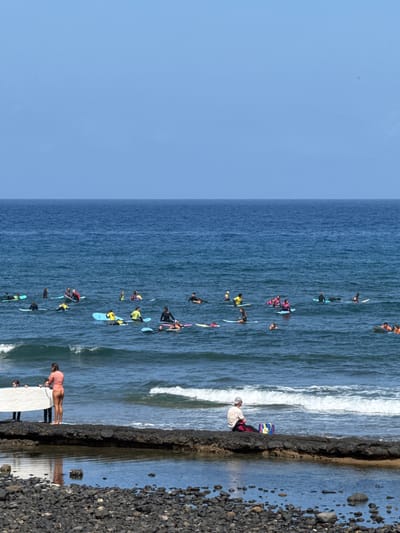 The image depicts a group of people on surfboards in the ocean, with a rocky shoreline to the right. The scene appears to be a surfing lesson or a gathering of surfers. The water is blue with some white waves breaking near the shore. The sky is clear and blue, suggesting a sunny day, likely during daylight hours.
There are many individuals scattered across the water, most of whom are sitting or kneeling on their surfboards. Some are actively paddling or appearing to be in the process of catching a wave. A few people are also visible on the rocky shore, one of them in the foreground wearing a striped shirt. There is a yellow and blue patterned board near the shore. The overall mood seems casual and recreational, consistent with a beach activity.
While specific details about time of day are not explicitly available, the bright sunlight indicates it is daytime. The presence of many people in the water suggests a popular spot for surfing, possibly a designated surfing area or a surf school in session. The location context provided is Arona, Spain. No visible text is present in the image.