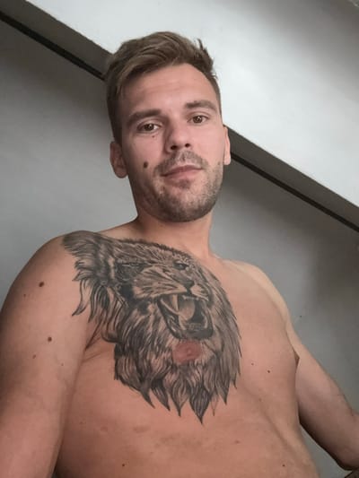 A close-up, low-angle shot shows a shirtless man from the chest up, facing the camera. He has short, light brown hair, a light beard, and a mole on his right cheek. A large tattoo of a roaring lion's head covers his left chest and shoulder. The background is a plain, light gray wall with a dark horizontal band across the top of the frame. The lighting appears to be natural, coming from the top right. No specific location in Sesto Calende, Italy, is discernible from the image.