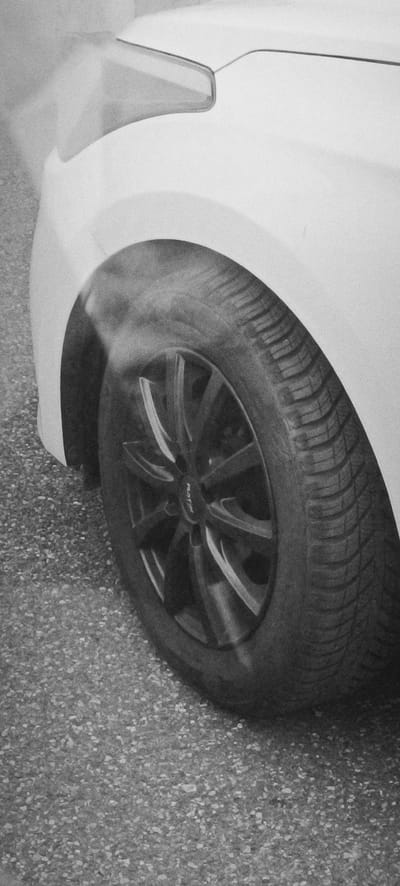 A black and white, close-up, eye-level shot shows the front passenger-side wheel and fender of a white car. The car is parked on a grey asphalt surface with visible aggregate. The tire is dark, with visible tread patterns, and appears to be slightly wet or steamy, as a light mist or vapor is rising from the top of the wheel well and obscuring part of the car's headlight. The wheel is a multi-spoke black alloy rim with some reflective highlights on the spokes. The car's fender is smooth and white, with a subtle curve leading to the headlight assembly. The background is out of focus, but appears to be an outdoor environment. No people are visible in the image. The scene is devoid of any distinguishing features that would allow for precise location identification within Waiblingen, Germany.