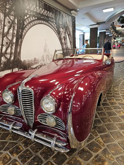 A high-angle shot shows the front of a maroon vintage car angled in front of a black and white backdrop of the Eiffel Tower. The car has a chrome grill and headlights and is parked on a cobblestone surface. In the background, people stand near a railing in a brightly lit museum setting. This image was taken in Архангельское, Russia.