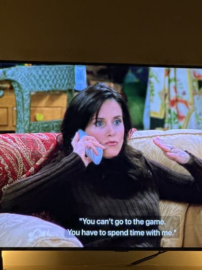 The image displays a television screen presenting a still frame from a fictional scene. The central figure is a female with dark hair and blue eyes, wearing a dark brown, ribbed turtleneck sweater. She holds a light blue cordless telephone receiver to her left ear with her left hand, and her right hand is raised mid-gesture, palm open and facing upward, as if making a point. Her expression is serious, with wide eyes and slightly parted lips. She is seated on a multi-patterned upholstered couch; the section to her left features a red, gold, and cream paisley design, while the section to her right is a plain cream or light gold.
In the background on the screen, to the left of the woman, is a section of green, intricately patterned furniture, possibly wicker or rattan, with a small, light-colored object inside one of its compartments. To her right, a colorful patterned fabric, likely a curtain or tapestry, with green, yellow, and possibly pink floral or abstract motifs is visible.
At the bottom of the screen, white subtitles are overlaid, reading: "You can't go to the game. You have to spend time with me."
The television screen itself has a narrow black border at the top and bottom. Above the screen, a plain light brown or beige wall is visible. Below the screen, a dark cable runs horizontally. The physical location where this television screen is present, such as Ericeira, Portugal, is not visually discernible within the provided image.