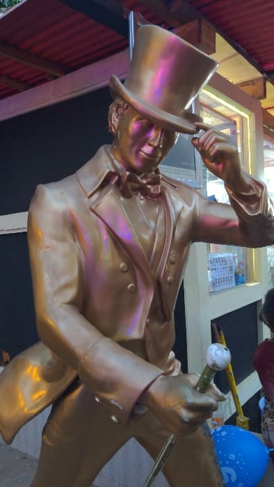 In Moshi, Tanzania, a golden-colored statue of a male figure is depicted in a semi-outdoor setting. The statue is dressed in a formal double-breasted suit with a bow tie and trousers, and wears a top hat, which he is tipping with his right hand. His left hand holds a walking stick topped with a spherical white knob. The statue's surface reflects light, showing iridescent purple and pink hues. The background features a red corrugated metal roof supported by wooden beams. To the right, a window or display case containing white items is visible, alongside a yellow pole leaning against a wall. A blue balloon with white markings is partially visible near the statue's base, and a portion of a person's arm in patterned clothing is present in the lower right background.