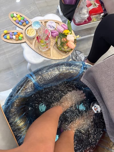 The image depicts an overhead view of a pedicure service in a nail salon in Naples, United States. A person's bare feet are submerged in bubbling water within a white pedicure basin, which is lined with a blue plastic bag. A light gray towel rests on the basin's edge to the right. To the left, a white stool supports a light-colored wooden tray shaped like a rabbit, featuring a spring or Easter theme. The tray holds two sections containing numerous small, colorful, speckled decorative eggs. Adjacent to these are three small white ceramic bowls containing different substances: a light blue gel, a yellow cream, and a purple cream. A clear glass pitcher with a stopper, filled with liquid and pink and yellow floral petals, stands in the center of the tray. To the right of the pitcher, a small woven nest-like basket contains artificial pink, purple, and yellow tulips, green foliage, and additional small speckled eggs. In the upper right background, another person's lower left leg, clad in black leggings and a gray and white athletic shoe, is visible. Behind this person, a white utility cart holds various salon supplies, including bottles, cotton pads in a jar, and a red box. The floor appears to be a light-colored tile.
