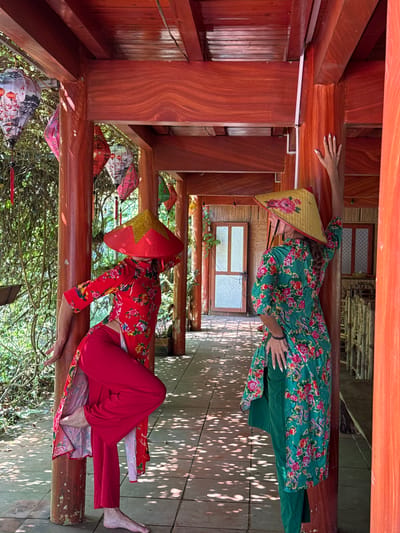 Two women, dressed in traditional Vietnamese ao dai and conical hats (nón lá), are depicted inside a wooden structure, likely a stilt house or viewing platform, in Thông Nguyên Commune, Vietnam. The image appears to be taken on a bright, sunny day, with dappled sunlight casting intricate shadows on the wooden surfaces.

One woman, on the left, wears a vibrant green ao dai adorned with a floral pattern of red and pink flowers and green leaves, paired with green pants. She also wears a yellow nón lá featuring a pink floral design. Her hand, with painted fingernails, rests casually on a wooden beam above her, while her other hand is on her waist.

The second woman, on the right, is dressed in a striking red ao dai with a similar floral motif, worn over red pants. Her red nón lá is embellished with a prominent gold star. She is seated on a wooden beam, with her legs extended and bare feet visible, and one hand resting on the beam beside her. Both women are posing in relaxed postures.

The surrounding environment includes numerous wooden beams and posts, creating an architectural frame. In the background, lush green foliage and trees are visible, along with windows featuring patterned grates. Decorative patterned lanterns hang in various spots. The scene captures a cultural moment in a natural, rustic setting. No visible text is present.