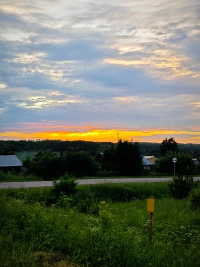 The image depicts a rural landscape at sunset, with a vibrant orange and yellow horizon visible above a dense line of trees. The sky is filled with scattered clouds, transitioning from lighter hues near the horizon to darker blues and grays higher up. In the foreground, a grassy field is dotted with wildflowers and taller plants. A dirt road or pathway runs horizontally across the middle ground, with several scattered buildings and trees visible beyond it. A yellow signpost stands prominently in the right foreground amidst the vegetation. The scene suggests a peaceful, natural setting, possibly on the outskirts of a rural community. The location is identified as Foki, Russia.