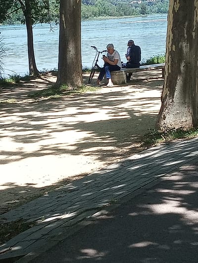 This is a medium shot, eye-level, outdoor photograph taken in Banyoles, Spain, of two individuals sitting on a bench near a lake. The foreground is dominated by the dappled shadows of trees cast upon a sandy path and a paved walkway. In the mid-ground, a woman in a white t-shirt and blue jeans is seated on a stone bench, looking at her phone. Next to her, a man in a dark shirt and cap is also seated on the bench, holding a phone. A bicycle with a black frame and orange accents is parked beside the woman. The background features a wide body of blue water, likely a lake, with trees lining the shore and buildings visible on the far side. The sun is bright, creating distinct shadows.