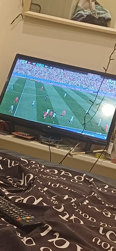 The image captures a close-up of a television screen displaying a sporting event, likely a rugby or American football match, given the on-screen graphics and player formations. The game is being played on a green field with white markings, and a stadium filled with spectators is visible on the left side of the screen. Several players, dressed in red and white uniforms, are scattered across the field.

On the right side of the image, a dark brown fabric with a white cursive script pattern is visible, partially obscuring the television. A remote control with several colored buttons rests on this fabric. The overall scene appears to be indoors, as indicated by the lighting and the presence of household items like a remote control.

Visible text on the television screen includes "LIVE" in the upper right corner, and on the bottom left, "33.35" and "103". The context of Stavroupoli Municipal Unit, Greece, suggests this is a domestic setting where the user is watching a live broadcast of a sporting event. No specific time of day or weather conditions can be determined from the image. The focus is on the screen content and the immediate surroundings.