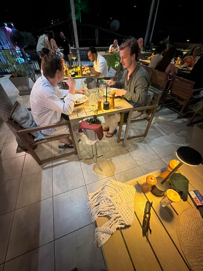 The image depicts a group of people enjoying a meal at an outdoor dining setting, likely in Ko Pha-ngan, Thailand. The scene is illuminated by artificial light, suggesting it is evening or night. The subjects are seated around a table, engaged in conversation and eating.
There are several individuals visible, including a man in a striped shirt in the foreground, another man in a dark shirt in the middle ground, and others further back. Objects on the table include plates with food, glasses, bottles, and a wine cooler. A lamp with wooden accents provides ambient light. A macrame wall hanging with fringe is placed on the table.
The setting appears to be a restaurant or a patio area, with tiled flooring and wooden furniture. Palm trees are visible in the background, hinting at a tropical location. The atmosphere seems relaxed and social. There is no indication of the weather, but the night setting implies it is likely warm.
A visible piece of text on a small rectangular object on the table reads "TEREA". The overall impression is one of a pleasant evening gathering with friends or family, enjoying food and company in a tropical environment.