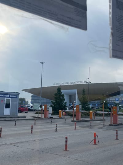 The image shows a view of the entrance to Bolshoye Savino Airport in Perm Krai, Russia. In the foreground, there is a paved area with several traffic cones and bollards. Beyond this, there is a parking lot with several cars visible. In the middle ground, there are automated entry gates for vehicles, complete with barriers and payment machines. To the left, a small grey building with the text "АДМИНИСТРАЦИЯ ПАРКОВКИ" (Parking Administration) is visible. In the background, the distinctive architecture of the airport terminal building can be seen, with the text "МЕЖДУНАРОДНЫЙ АЭРОПОРТ ПЕРМЬ" (Perm International Airport) illuminated on its facade. The sky is overcast, and the overall lighting suggests it is daytime.