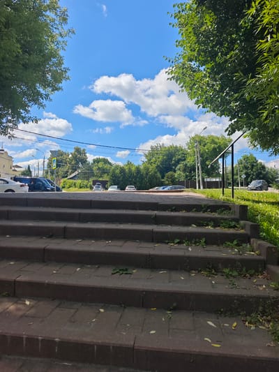 The image captures a view from the bottom of a brick staircase, looking upwards towards a street scene on a sunny day with a bright blue sky and scattered white clouds. The staircase, made of reddish-brown brick pavers, is slightly overgrown with small weeds and has fallen leaves on its steps. A black metal handrail is visible on the right side of the staircase, extending upwards and to the right.
Beyond the staircase, the scene unfolds with a road lined by green trees. Several cars are parked or driving on the road, including a white car, a black van with people near its open doors, and other vehicles further down. A pedestrian crossing sign is visible on the left side of the road, near a light-colored building with a cross on its roof, suggesting a religious establishment. Streetlights are also present. The background is filled with lush green trees and foliage, indicating a suburban or park-like setting within the city of Chekhov, Russia. The overall impression is a typical urban street scene with natural elements.