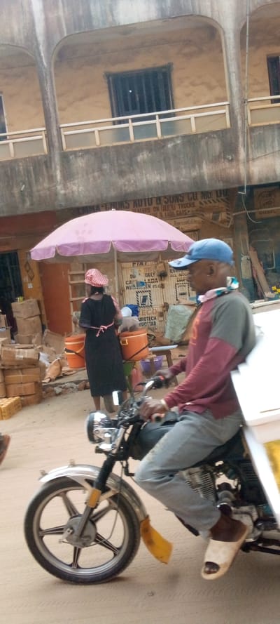 This image depicts a daytime street scene in Nnewi, Nigeria, featuring a man operating a motorcycle-tricycle and a woman standing beneath a large umbrella.
In the foreground, a dark-skinned male is seated on and operating a motorcycle-tricycle, moving towards the right side of the frame. He wears a blue baseball cap, a two-toned long-sleeved shirt (maroon sleeves, grey torso), light blue jeans, and light-colored slip-on sandals. A patterned cloth is tied around his neck, and he has a small earring in his left ear. The motorcycle's front is visible, including a chrome headlight, a chrome front fender with a yellow mud flap, and a five-spoke alloy wheel. A white cargo box or sidecar structure is partially visible to the right of the rider.
In the midground, a woman stands with her back to the viewer, positioned under a large, open pink umbrella. She wears a dark-colored dress with a pink/red tie at the waist and a red-and-white checkered head covering. Behind her, two orange buckets rest on a small wooden stool. To the left of the woman, stacks of cardboard boxes are piled, and a large sack is visible.
The background features a two-story building with a light brown facade and concrete accents, exhibiting signs of wear and weathering. The ground floor displays various commercial signs, including "EMMA-LADURA AUTO & SONS CO. NIG. LTD. DEALERS ON HINTI TRUCKS FIAT & LADA CARS" and "FIAT DAILY CA LADA WINSCHEN etc. EUROTECH UPSTAR." The upper floor has arched architectural supports, windows with dark grilles or shutters, and a balcony with light-colored railings. The street surface is composed of packed dirt or dust.