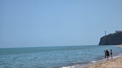 The image displays a coastal scene in Byala, Bulgaria, under a clear, bright blue sky. A vast expanse of calm, blue-green sea occupies the mid-ground, meeting a sandy beach in the foreground. The horizon line is flat and extends across the frame.

On the right side of the image, a dark, rugged cliff rises sharply from the waterline, topped by sparse green vegetation. A tall, slender metal lattice tower stands prominently near the cliff's edge, with a white-roofed building partially visible behind it further inland.

Near the shoreline in the lower right, three individuals are present. One person, wearing dark shorts and a light cap, walks on the wet sand towards the water, facing away from the camera. Two other individuals, appearing to be women in swimsuits, stand in the shallow water beside the first person, observing the sea. Gentle waves break softly on the shore, creating white foam. Small dark specks, possibly birds, are visible against the blue sky, with one more distinct bird flying closer to the horizon on the right.