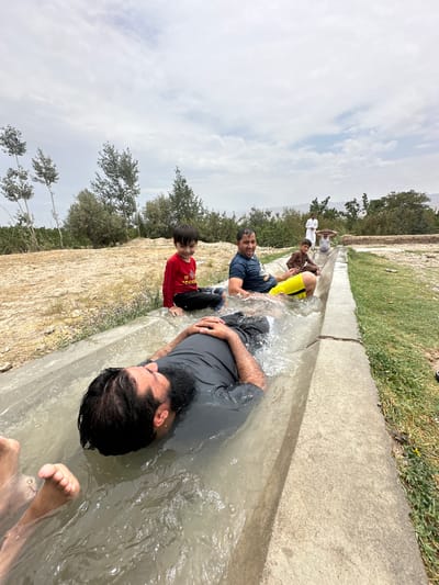 In Kan Mehtarzai, Pakistan, a group of people are enjoying a water channel. The foreground features a man lying on his back in a concrete water channel filled with murky water, his head submerged. His legs and feet are visible above the water. To his left, another man is also partially submerged in the channel, wearing a dark t-shirt and yellow shorts, with a broad smile on his face. A young boy in a red long-sleeved shirt and black pants sits on the edge of the channel, smiling. Further back, a man in a brown outfit sits in the channel, and behind him, two men stand in the distance, one in white traditional attire. The environment consists of a dry, sandy ground with sparse vegetation and trees, and a cloudy sky. The scene appears to be an informal recreational activity, likely during warm weather, utilizing the water channel for cooling off.