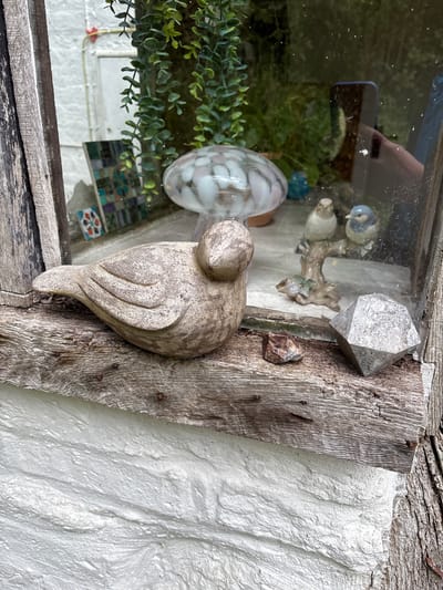 The image presents a detail of an outdoor decorative arrangement. Dominating the view is a weathered wooden frame, secured to a textured white wall. Affixed to the wooden frame are several objects. A carved stone bird figure perches prominently on the frame. A crystalline structure is also attached, alongside a smaller stone fragment.
Behind the frame, a glass surface reflects a scene with greenery and other decorative elements, including a spotted glass ornament, and a porcelain bird figurine, hinting at an indoor or sheltered space. The reflected environment adds depth and visual complexity to the composition.
This image was taken in Pulverbatch, United Kingdom.
