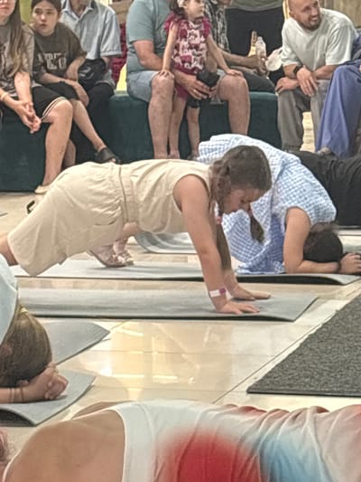 In Tri Protoka, Russia, a group of people are engaged in a yoga session, with several individuals positioned on yoga mats on the floor. In the foreground, a young woman with braided hair is in a plank position, wearing a light-colored jumpsuit and a purple and white wristband. To her right, another person in a blue and white checkered shirt is also on their mat, resting their head on their hands. Further back, a child in a patterned dress sits between adults. The background shows more individuals seated and standing, with a dark green couch visible. The flooring appears to be a combination of tiled and carpeted surfaces.