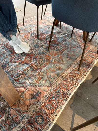 A high-angle shot depicts a portion of a room. A dark blue chair sits on the left side of the frame, with its legs extending onto an intricate area rug. The rug features a blend of blue, brown, and rust-colored patterns. A pair of white socks worn with dark blue jeans are visible in the lower-left corner of the image. The room appears to have a hard floor, possibly tile or concrete. The image does not give enough information to determine if the photo was taken in Chkalov, Armenia.